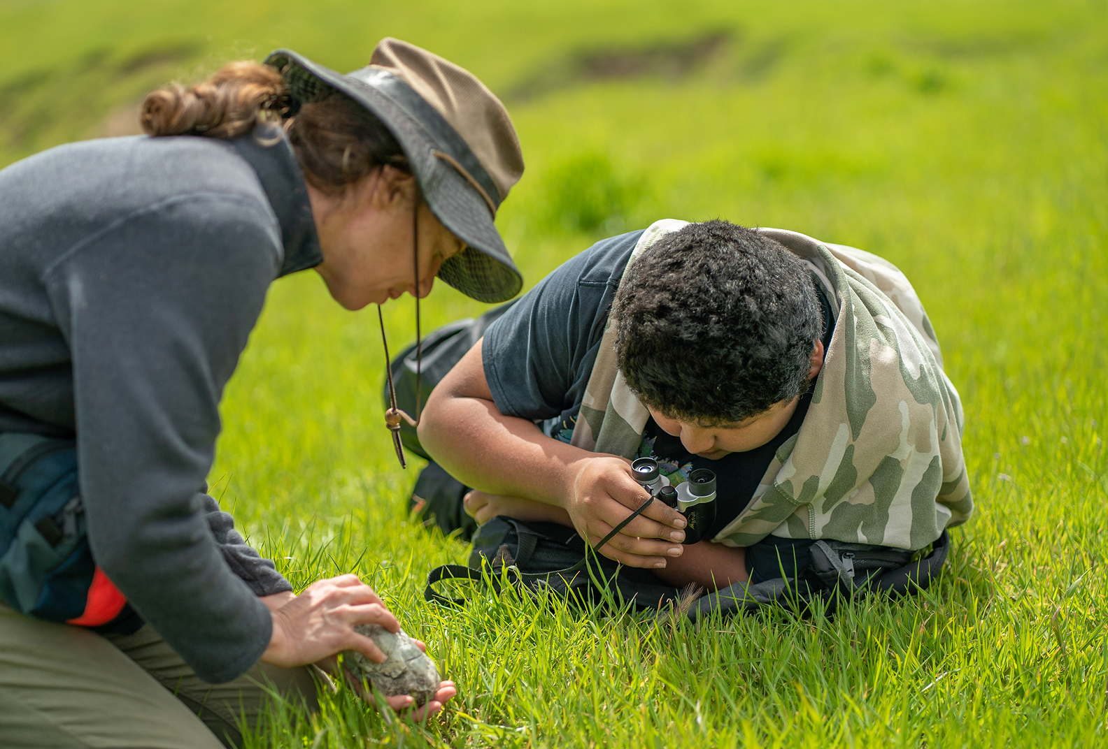 Student and teacher crouch to inspect a rock in a meadow