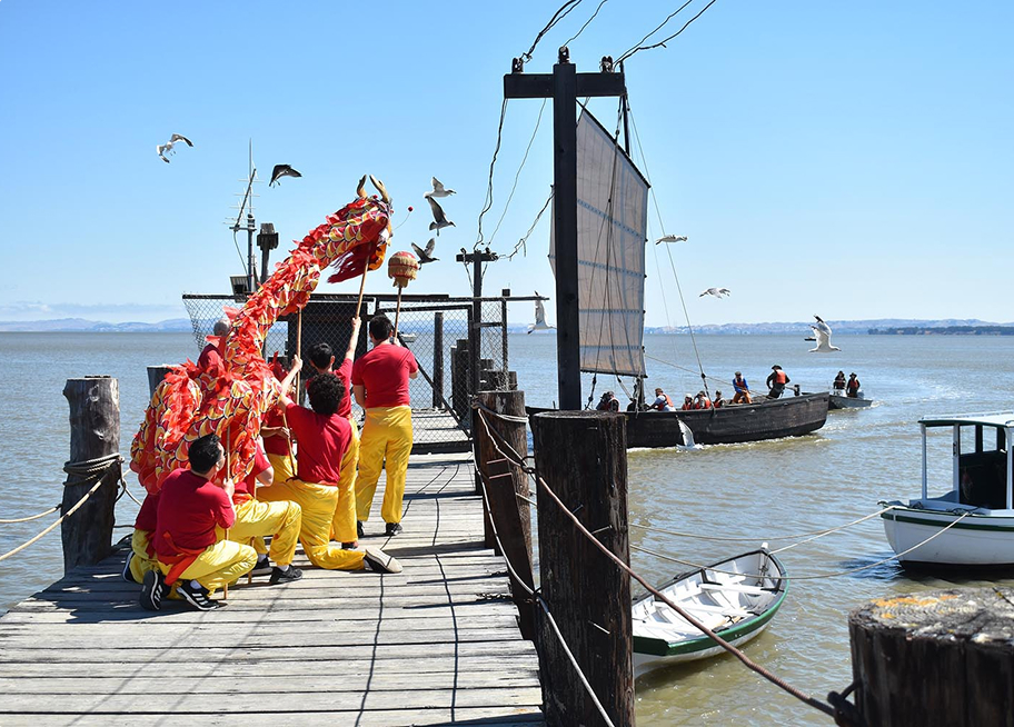 Chinese dragon puppet with performers on a pier with boats in the background.